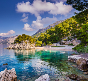 Felsige Küste mit türkisfarbenem Wasser und Pinienbäumen in Kroatien, im Hintergrund Berge und ein kleiner Strand mit Booten.