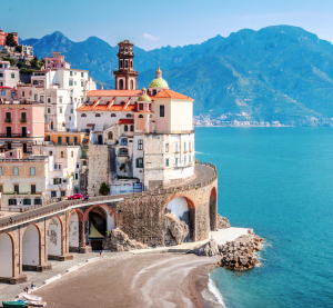 Küstenstadt Amalfi in Italien mit bunten Häusern auf einer Klippe, darunter eine Strandbucht und das türkisfarbene Mittelmeer, im Hintergrund Berge.