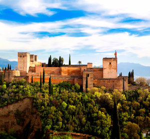 Die Alhambra in Granada, Spanien, umgeben von üppigem Grün, mit den schneebedeckten Bergen der Sierra Nevada im Hintergrund unter blauem Himmel.