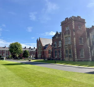 Historisches Gebäude der Queen’s University Belfast mit Rasenfläche und blauem Himmel im Hintergrund.