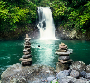 Wasserfall, der in einen klaren Naturpool st&uuml;rzt, flankiert von gestapelten Steinen im Vordergrund.