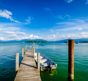 Holzsteg mit Boot auf einem ruhigen See in Bayern, im Hintergrund eine kleine Insel und die Alpen unter blauem Himmel mit weißen Wolken.