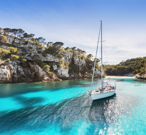 Segelboot in einer türkisblauen Bucht auf Menorca, umgeben von steilen Felsklippen mit Pinienbewuchs und klarem Himmel darüber.