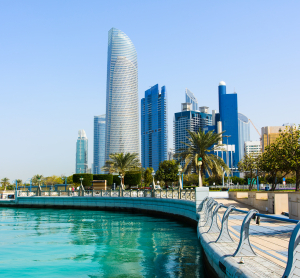 Moderne Skyline von Abu Dhabi mit hohen Glasgebäuden am türkisfarbenen Wasser, gesäumt von einer Promenade mit Palmen und Geländern.