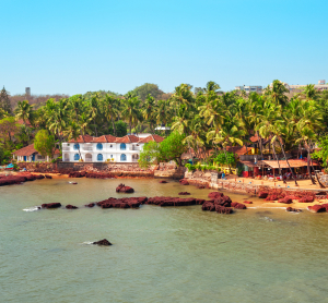 Blick auf eine Küstenlandschaft in Goa mit weißen und roten Kolonialgebäuden, gesäumt von zahlreichen Palmen am ruhigen Meer.