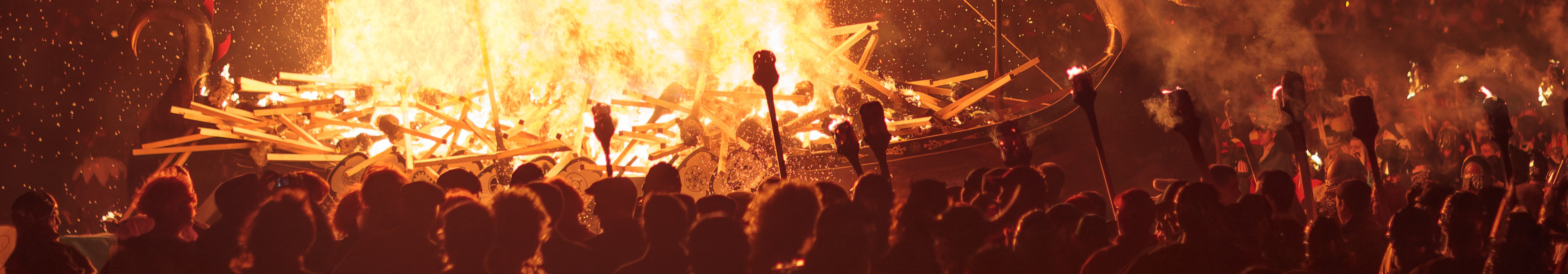 Ein großes, brennendes Wikingerschiff bei einem Feuerfest in der Nacht, mit vielen Zuschauern und Fackeln.