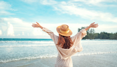 Frau mit Strohhut und ausgestreckten Armen genießt ihren Urlaub am tropischen Sandstrand mit Blick aufs Meer