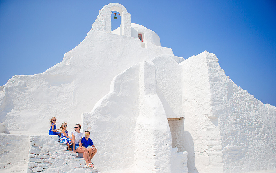 Familie sitzt auf den Treppen neben einer ikonischen weißen Kirche auf Mykonos, umgeben von strahlend blauem HImmel. 