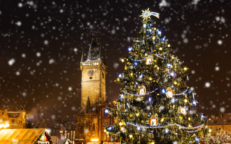 Festlich beleuchteter Weihnachtsbaum auf dem Weihnachtsmarkt in Prag, einem der schönsten Adventsmärkte weltweit, mit dem historischen Uhrturm im Hintergrund und sanft fallendem Schnee.