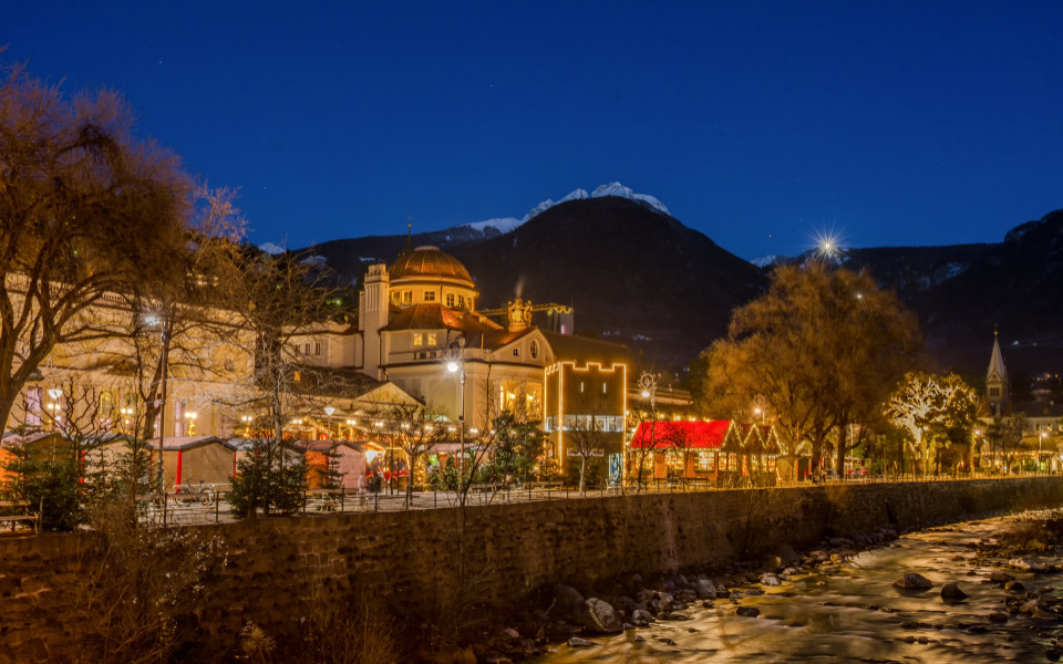 Romantischer Christkindlmarkt in Meran, Südtirol, mit festlich beleuchteten Ständen, winterlicher Bergkulisse und stimmungsvoller Atmosphäre entlang des Flusses.