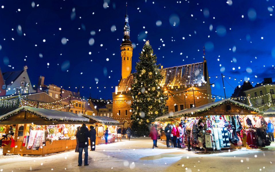 Winterlicher Weihnachtsmarkt in Tallinn mit festlich geschmückten Holzhütten, leuchtendem Weihnachtsbaum und Schneeflocken vor der historischen Rathauskulisse.