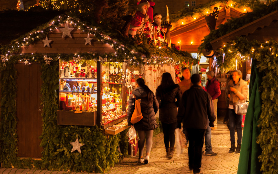 Stimmungsvoll beleuchtete Weihnachtsmarktstände in Paris, geschmückt mit Tannenzweigen und Lichterketten, während Besucher festliche Dekorationen und handgefertigte Geschenke entdecken.