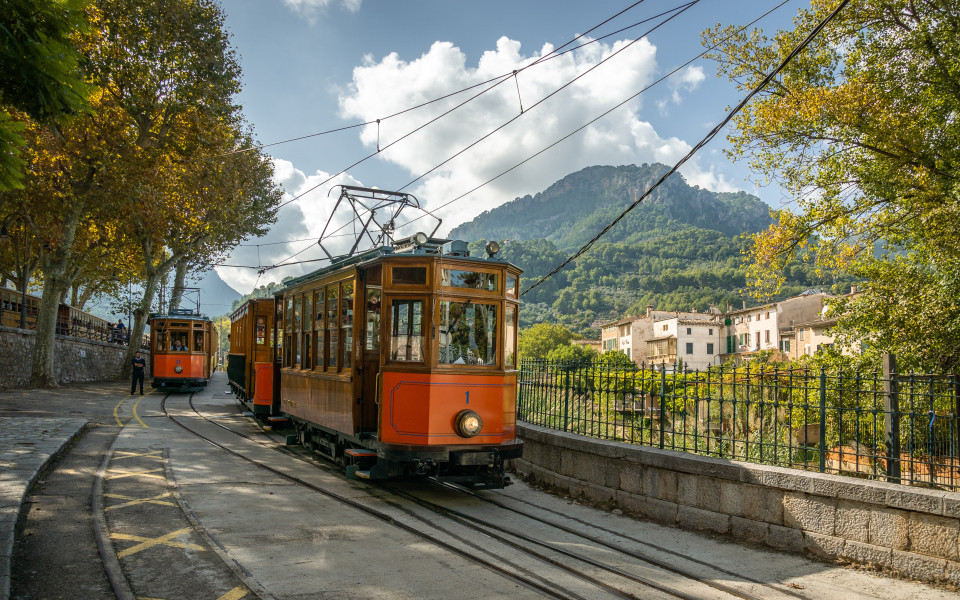 Historische Straßenbahn „Roter Blitz“ auf Mallorca, umgeben von grünen Bergen, malerischen Dörfern und mediterraner Landschaft.