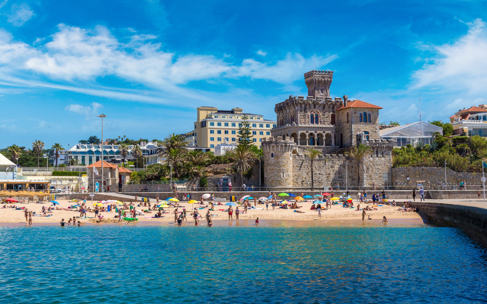 Sonniger Strand in Estoril mit goldenem Sand, türkisblauem Wasser und dem markanten Castelo dos Mouros im Hintergrund.