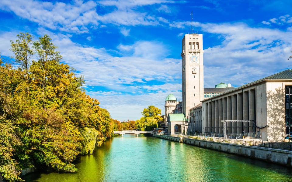 Sehenswürdigkeit in München: Das Deutsche Museum in München, malerisch am Ufer der Isar gelegen, umgeben von herbstlich gefärbten Bäumen und strahlend blauem Himmel.