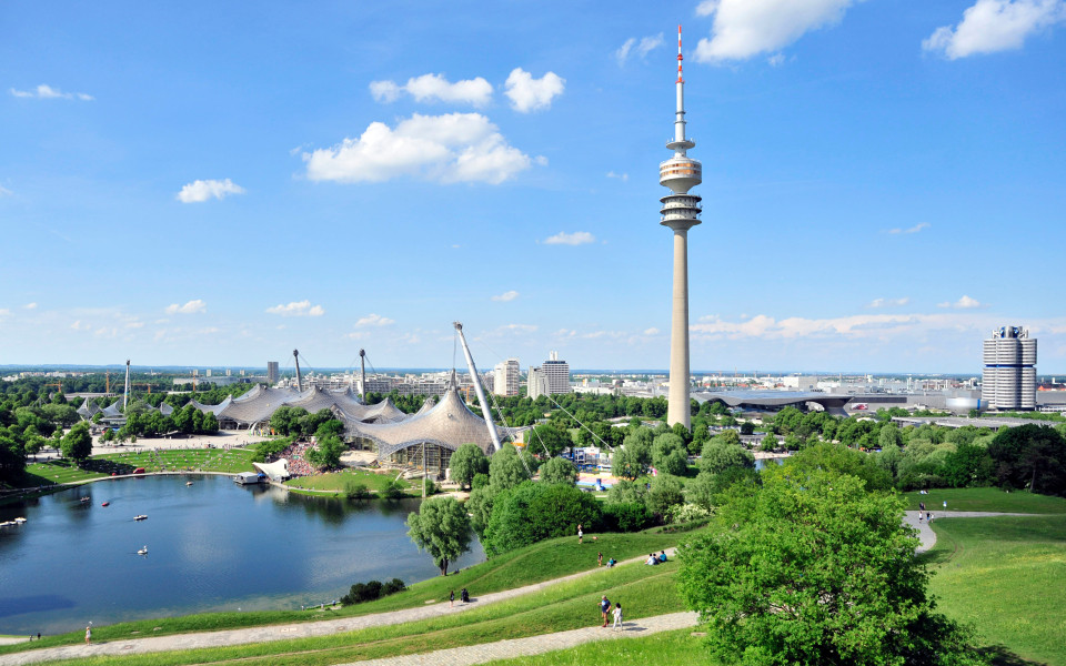 Geheimtipp in München: Der Olympiapark in München mit dem markanten Olympiaturm, der ikonischen Zeltdacharchitektur und dem idyllischen See, umgeben von grünen Wiesen unter blauem Himmel.