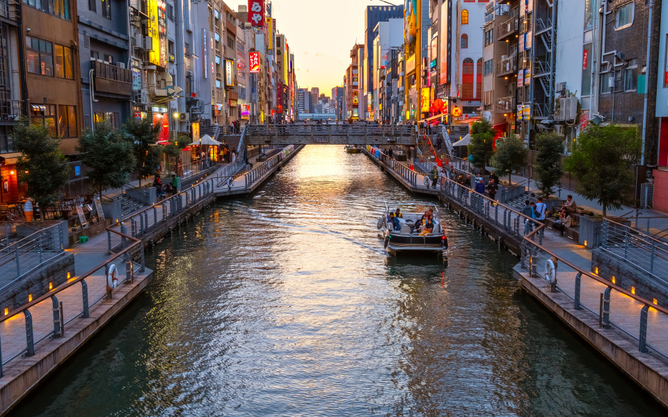 Belebter Kanal Dotonbori in Osaka mit Boot und beleuchteten Häuserfassaden bei Sonnenuntergang.