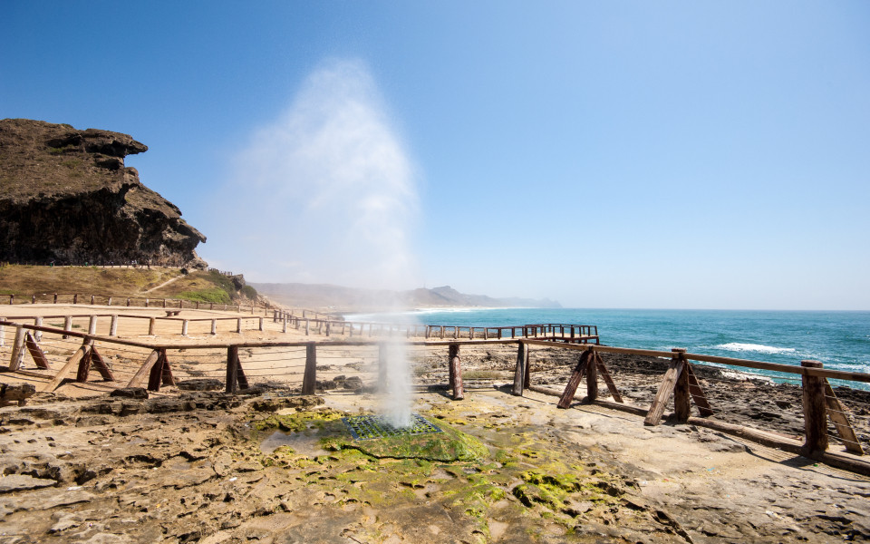 Al Mughsail Beach in Oman mit einem Wassergeysir, der aus den Felsen schießt, vor dem weiten Ozean und einer Holzpromenade.