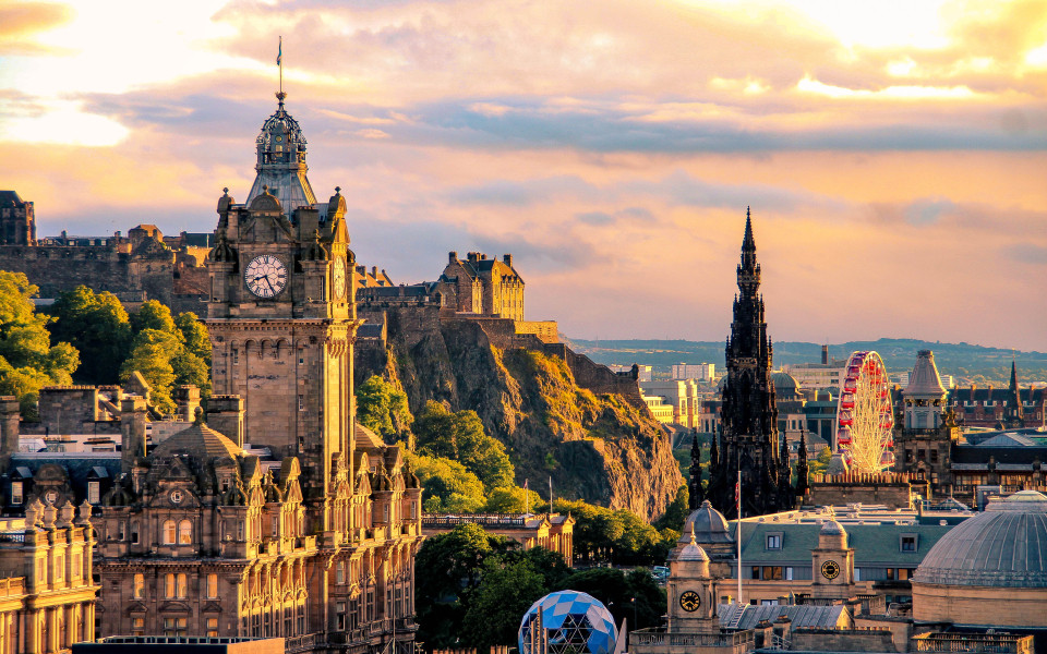 Panoramablick auf Edinburgh bei Sonnenuntergang mit dem markanten Uhrturm des Balmoral Hotels, dem Edinburgh Castle auf dem Hügel, dem gotischen Scott Monument und einem Riesenrad im Stadtzentrum.