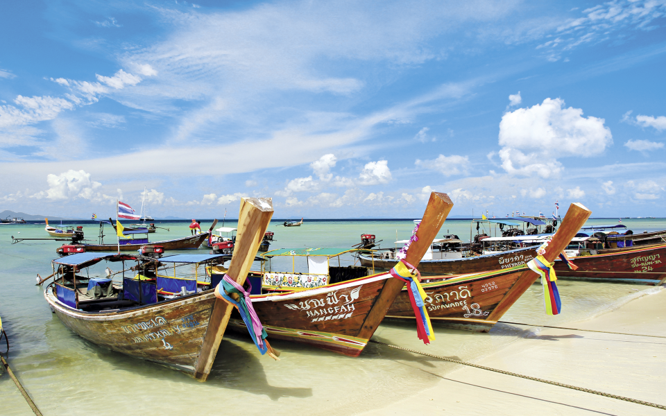 Traditionelle Longtail-Boote in Thailand am weißen Sandstrand von Phuket, verziert mit bunten Bändern, vor türkisfarbenem Wasser und blauem Himmel mit Wolken.