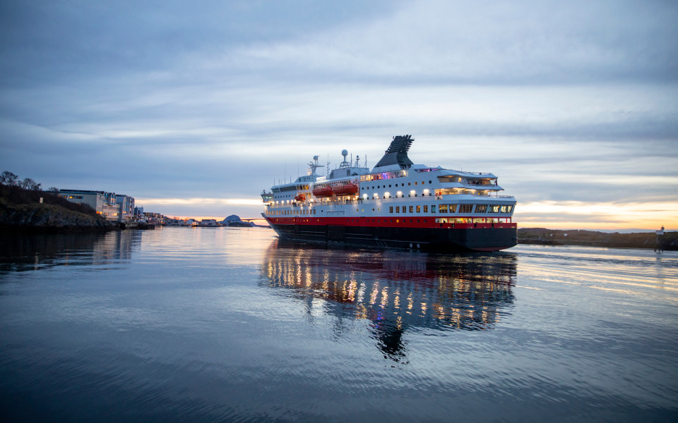 Ein Schiff der Hurtigruten fährt bei Abenddämmerung durch einen ruhigen Fjord in Norwegen. 