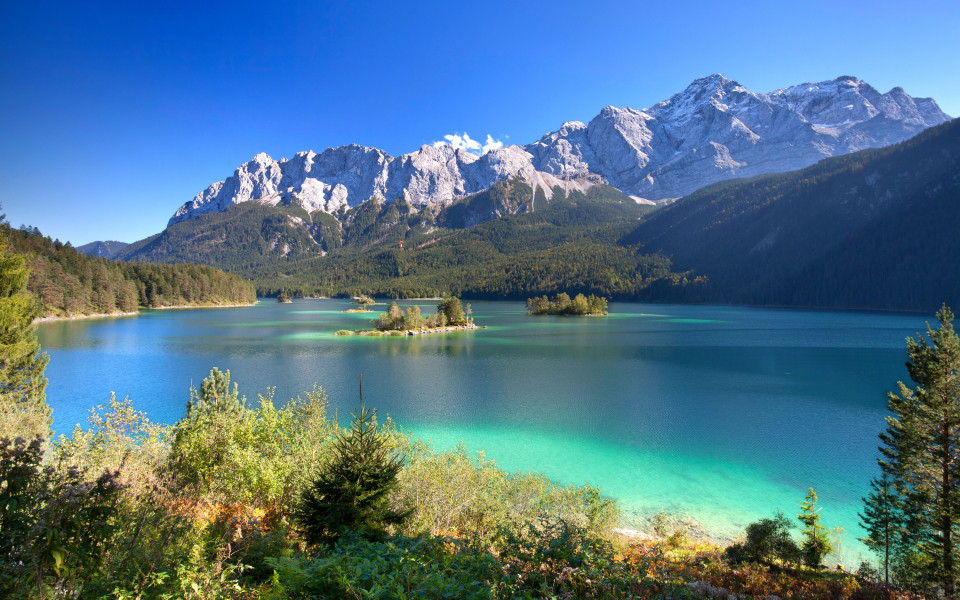 Blick auf den türkisfarbenen Eibsee mit kleinen Inseln, umgeben von dichtem Wald und den schneebedeckten Gipfeln der Zugspitze in Bayern, Deutschland.        ChatGPT fragen